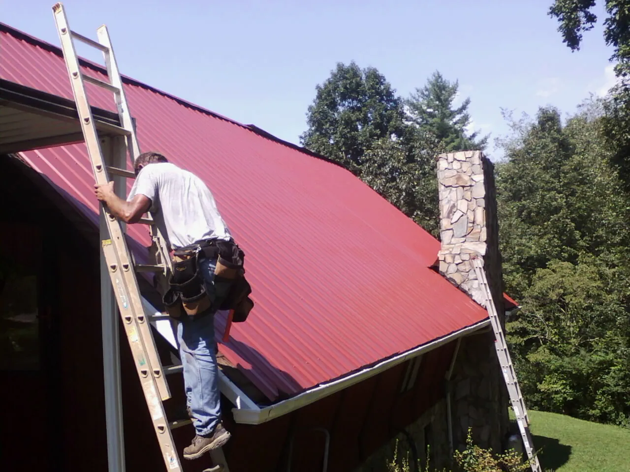 Roofer on a ladder beside a red metal roof and stone chimney — Soffit & Fascia Repair by Roofing Brothers in Stafford